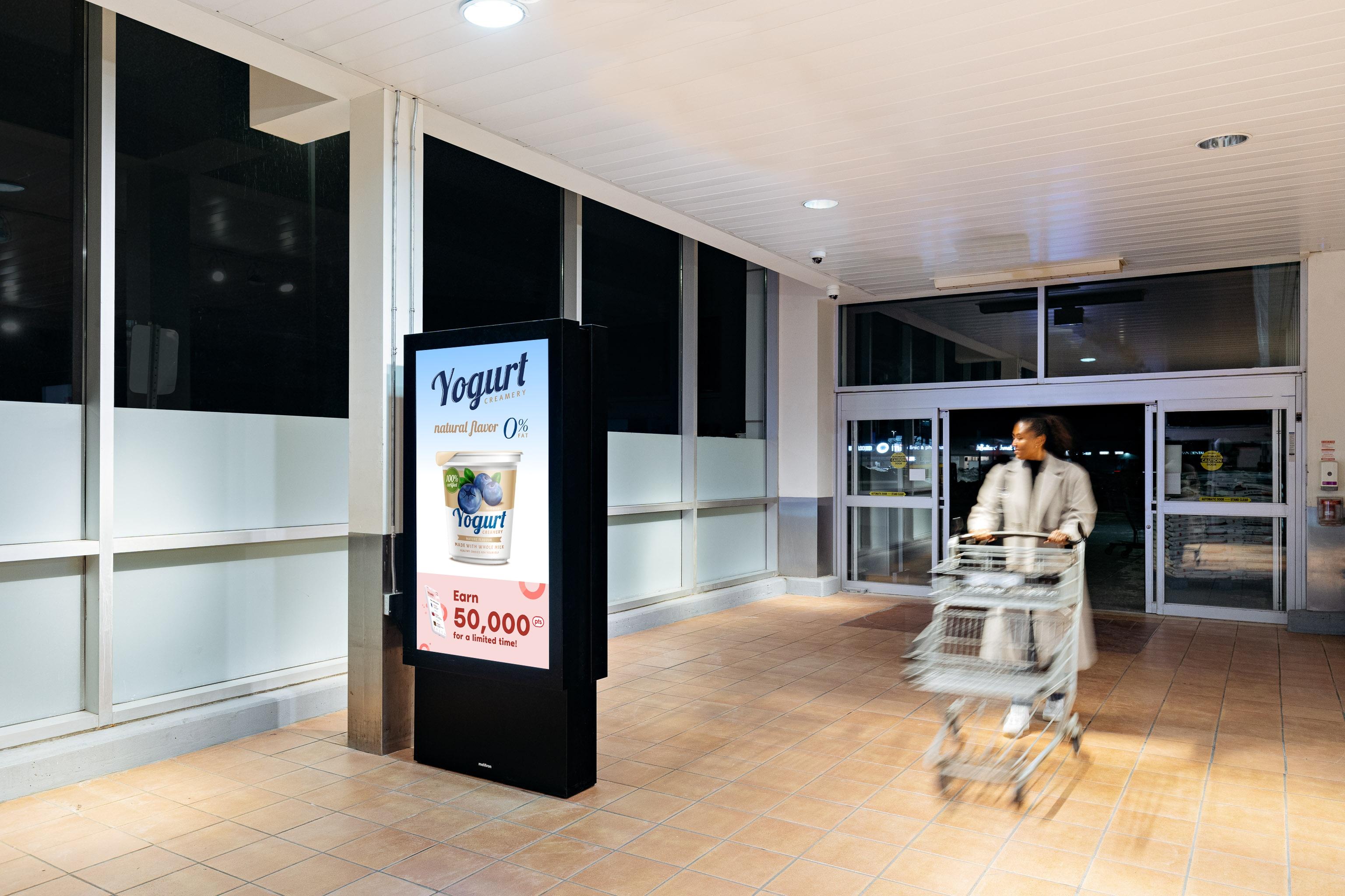 A woman walks by a screen at the front entrance of a grocery store with a yogurt advertisement on it.
